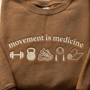 May include: Brown crewneck sweatshirt with the embroidered text "movement is medicine" and white embroidered icons of a dumbbell, kettlebell, running shoe, jump rope, and yoga mat.