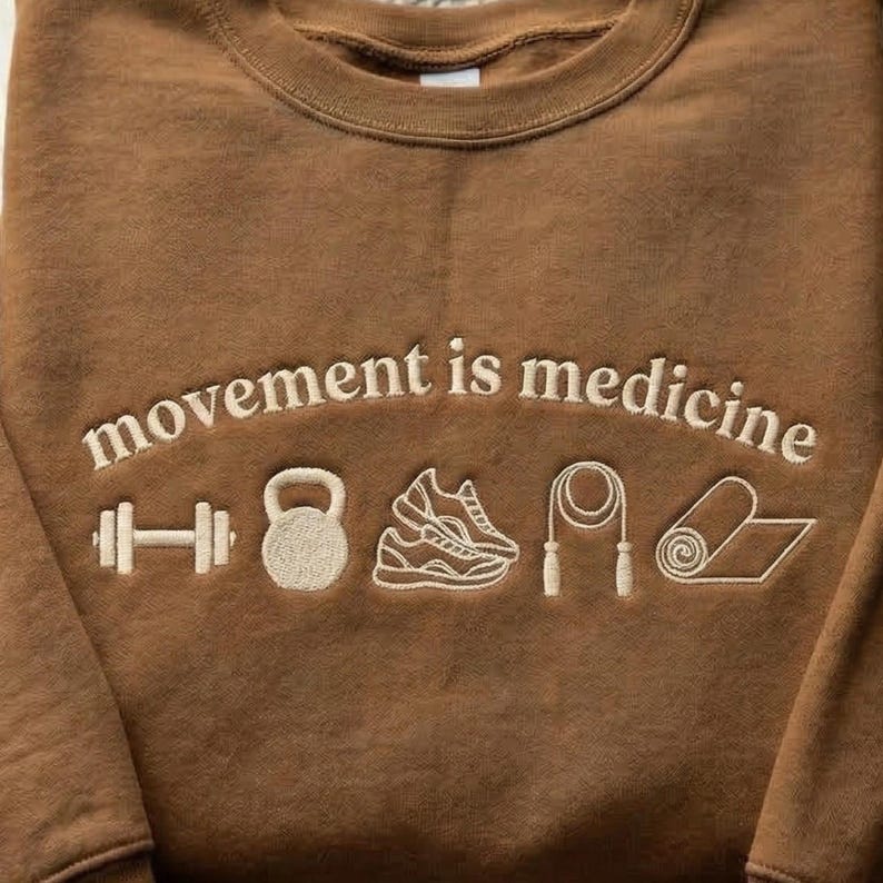 May include: Brown crewneck sweatshirt with the embroidered text "movement is medicine" and white embroidered icons of a dumbbell, kettlebell, running shoe, jump rope, and yoga mat.