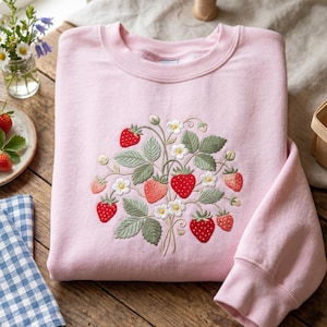 May include: A light pink crewneck sweatshirt featuring a central embroidered design of a strawberry plant with red berries, green leaves, and small white flowers. The embroidery is detailed with various shades of green, red, and white thread.