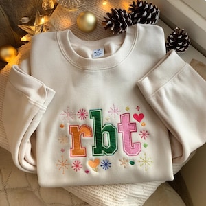 May include: Cream-colored sweatshirt with embroidered "rbt" letters in orange, green, and pink, surrounded by snowflakes, hearts, and stars. The long-sleeved top is made of a soft material.