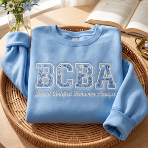May include: A light blue sweatshirt with the embroidered letters "BCBA" in white, adorned with floral designs. Below the letters, the text reads "Board Certified Behavior Analyst". The sweatshirt is folded on a woven tray.