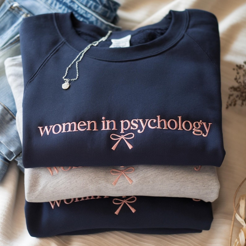 May include: A stack of three sweatshirts, navy blue and gray, embroidered with "women in psychology" in pink script. Each sweatshirt has a pink bow graphic below the text. A silver necklace with a small pendant rests on the top sweatshirt.