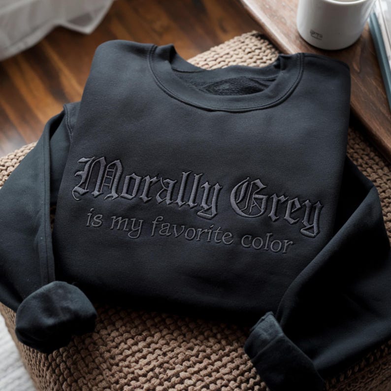 Peut inclure: Un sweat-shirt noir avec la phrase brod&eacute;e "Morally Grey is my favorite color" en police gothique. Le sweat-shirt est pli&eacute; sur une surface tiss&eacute;e, avec les manches retrouss&eacute;es. Une tasse blanche est visible en arri&egrave;re-plan.