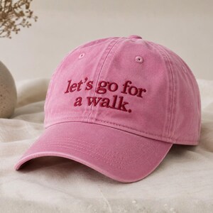 May include: A pink baseball cap with the embroidered text "let's go for a walk." The cap features a curved peak and a classic six-panel design. The text is in a darker shade of pink.