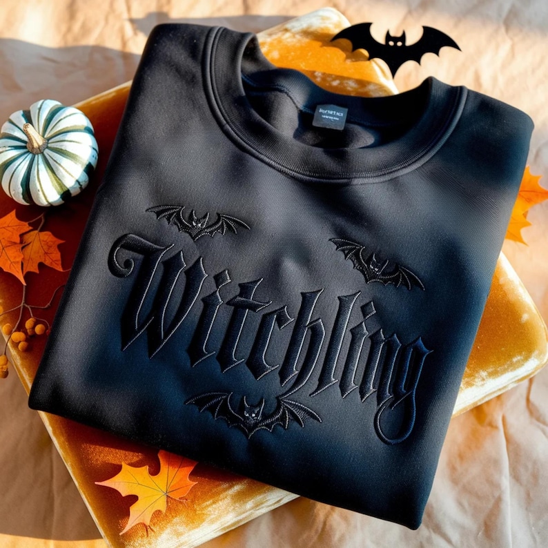 May include: Black sweatshirt with the word "Witchling" embroidered in a gothic font, accompanied by bat designs. The sweatshirt is displayed on a gold velvet surface, with a small pumpkin and autumn leaves nearby.