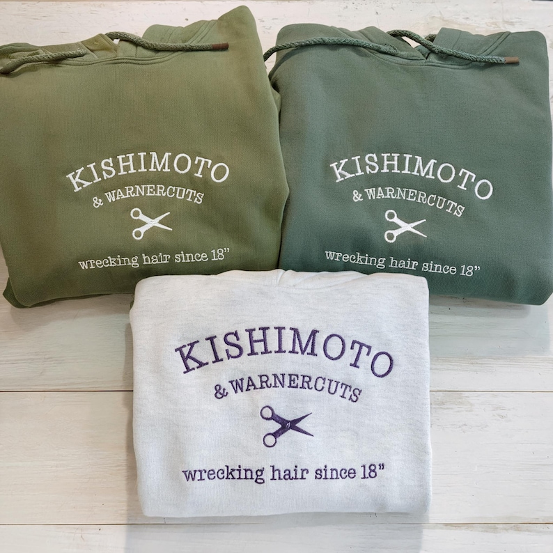 May include: Three hooded sweatshirts in olive green and light gray. Each sweatshirt has the embroidered text "KISHIMOTO & WARNERCUTS" with scissors and the phrase "wrecking hair since 18".