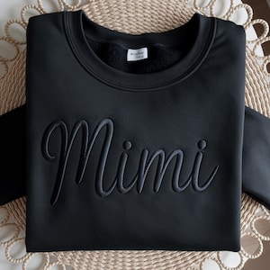 以下が含まれることがあります： 黒色のスウェットシャツ。筆記体で「Mimi」の刺繍が施されています。柔らかい素材でできており、クルーネックです。写真は真上から撮影されています。