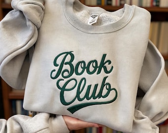 Puede incluir: Una sudadera gris claro con las palabras "Book Club" bordadas en guion verde oscuro. La sudadera tiene cuello redondo y mangas largas. El fondo muestra una estantería.