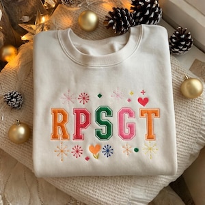 May include: Cream-colored sweatshirt with embroidered letters "RPSG T" in orange, pink, and green. The design includes snowflakes, hearts, and other colorful accents. The sweatshirt is displayed on a soft, textured surface with holiday decorations.