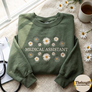 Puede incluir: Una sudadera de cuello redondo color verde oliva con margaritas blancas bordadas de centros amarillos que rodean el texto MEDICAL ASSISTANT en una fuente sans-serif. El diseño está centrado en el pecho de la prenda de manga larga.