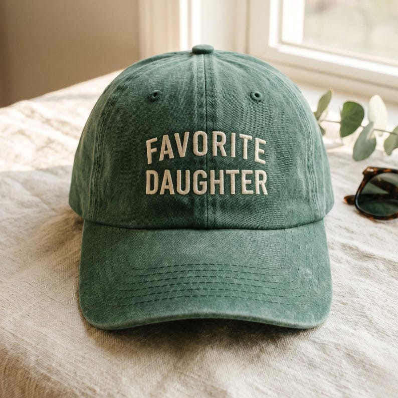 May include: A green baseball cap with the words "FAVORITE DAUGHTER" embroidered in white. The cap is made of a soft, textured fabric and has a curved brim. Sunglasses are visible in the background.