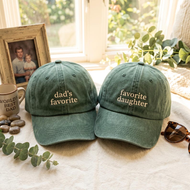 May include: Two green baseball caps with white embroidered text. One says "dad's favorite" and the other says "favorite daughter." The caps are on a white surface, with a framed photo and a mug in the background.