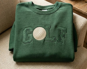 May include: A dark green sweatshirt with the word "GOLF" embroidered in a matching green thread. A golf ball is embroidered in the center of the word. The sweatshirt is folded and resting on a beige upholstered chair.