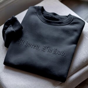 May include: A dark grey sweatshirt featuring the embossed phrase "Hi bored. I'm Dad." The folded sweatshirt is placed on a light grey surface. The sleeve is folded over, showing the ribbed cuff.