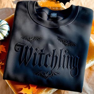 May include: Black sweatshirt with the word "Witchling" embroidered in gothic lettering, along with three bat designs. The sweatshirt is displayed on a velvet surface with autumn-themed decorations.