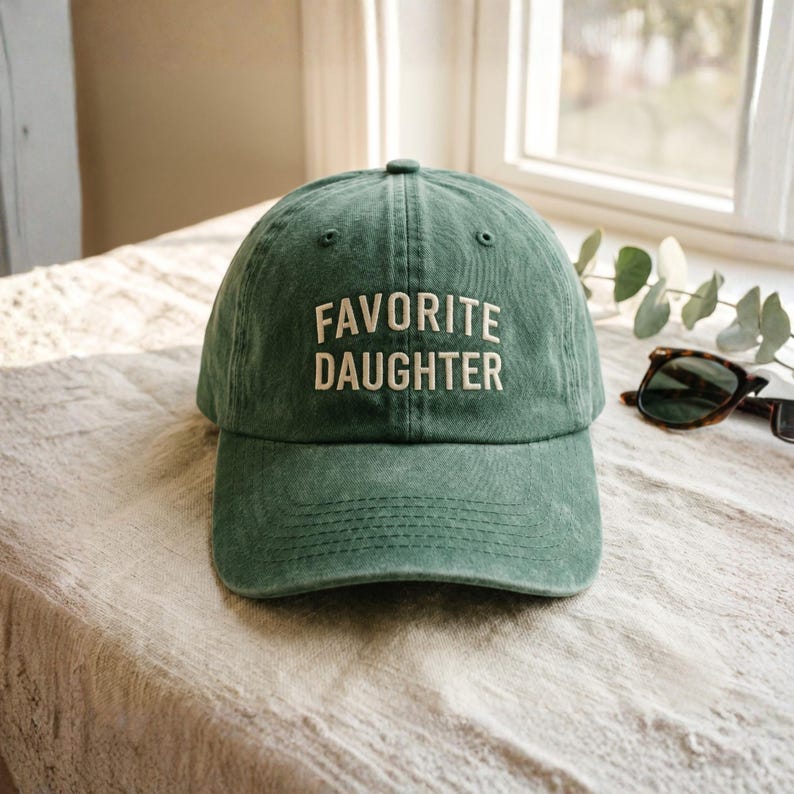 May include: A green baseball cap with the text "FAVORITE DAUGHTER" in white. The cap is made of a soft material and has a curved brim. The hat is sitting on a beige surface.