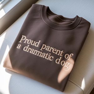 May include: A folded brown sweatshirt with the embroidered text "Proud parent of a dramatic dog" in a light beige color. The sweatshirt has a crew neck and is made of a soft material.