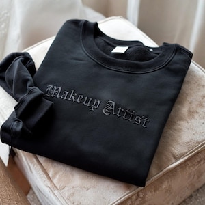 May include: Black sweatshirt with the embroidered text "Makeup Artist" in a gothic font. The sweatshirt is folded neatly on a beige upholstered chair. The sleeves are folded over the chair.