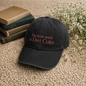 Könnte beinhalten: Schwarze Baseballkappe mit dem gestickten Text "The Bride needs a Diet Coke" in Rosa. Die Kappe liegt auf einer strukturierten Oberfläche, im Hintergrund sind Bücher und Blumen zu sehen.