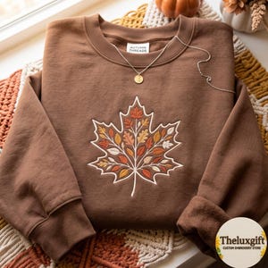 Puede incluir: Una sudadera marrón de cuello redondo con un gran diseño de hoja de arce bordado, compuesto por hojas más pequeñas en tonos naranja, amarillo y marrón. La etiqueta interior del cuello dice AUTUMN THREADS. Un collar dorado descansa sobre el escote.