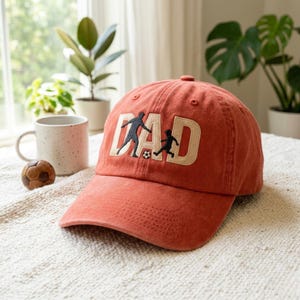 May include: A coral-colored baseball cap with the word "DAD" embroidered in cream-colored letters. The "A" is formed by silhouettes of a person and a child playing soccer. A soccer ball is also embroidered on the cap.