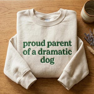 May include: Beige sweatshirt with the embroidered text "proud parent of a dramatic dog" in green. The sweatshirt has a crew neck and long sleeves. The image is taken from a top-down perspective.