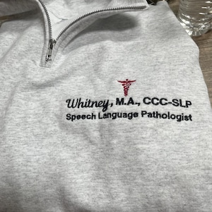 May include: A light gray sweatshirt with a quarter-zip neckline. The sweatshirt features embroidered text that reads "Whitney, M.A., CCC-SLP Speech Language Pathologist" below a red medical symbol.