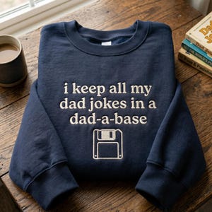 Könnte beinhalten: Marineblaues Sweatshirt mit dem gestickten Schriftzug "i keep all my dad jokes in a dad-a-base" und einer Diskettengrafik. Das Kleidungsstück liegt auf einem Holztisch, mit einer Tasse und Büchern im Hintergrund.