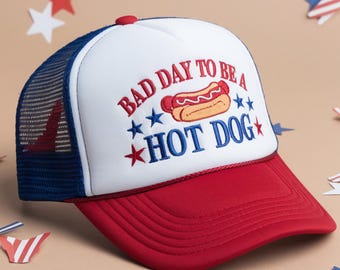 May include: A red, white, and blue trucker hat with the text "BAD DAY TO BE A HOT DOG" embroidered on the front. The hat features a red brim, a white front panel with a hot dog graphic, and a blue mesh back.