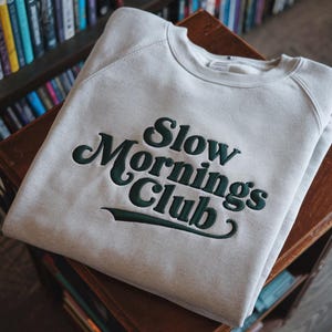 May include: A pale grey sweatshirt featuring the embroidered words "Slow Mornings Club" in dark green. The garment is folded on a wooden surface, with a bookshelf in the background.