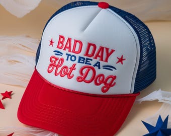 May include: A red, white, and blue trucker hat with the text "BAD DAY TO BE A Hot Dog" embroidered on the front. The hat has a red brim, a white front panel, and a blue mesh back. A red button is on top.