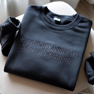 May include: Black sweatshirt with the embroidered text "Somebody's Bomb Ass Massage Therapist." The crew neck sweatshirt is made of a soft material and has ribbed cuffs. The sweatshirt is folded on a light-colored surface.
