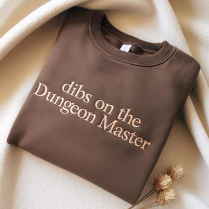 May include: A brown sweatshirt with the embroidered text "dibs on the Dungeon Master" in a light tan color. The sweatshirt is folded and resting on a cream-colored fabric. Dried flowers are in the bottom right corner.