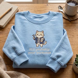 May include: A light blue crewneck sweatshirt featuring a graphic of a cartoon cat dressed as a doctor, holding a medical tool and a clipboard. Below the cat, the text reads, "it's probably your hormones" in a simple, white, sans-serif font.