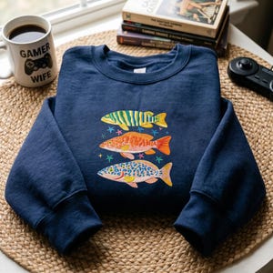 May include: Navy blue sweatshirt featuring embroidered fish in shades of green, orange, and blue. The design includes star accents. A white mug with the text "GAMER WIFE" and a game controller graphic is visible.