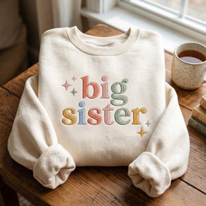 May include: Cream-coloured sweatshirt with the embroidered text "big sister" in colourful letters. The letters are in shades of red, green, yellow, and blue, with star accents. The sleeves are cuffed. A mug of tea and books are in the background.