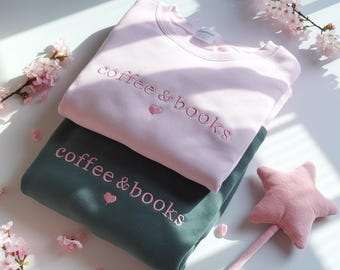 May include: Two sweatshirts, one light pink and one dark green, are stacked. The light pink sweatshirt has the embroidered text "coffee & books" with a heart. The dark green sweatshirt has the same text and heart. A pink star wand is on the right.