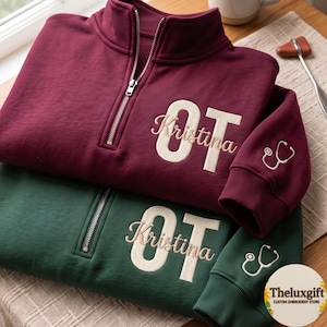 Puede incluir: Una sudadera color granate y otra verde oscuro con cremallera de cuarto, bordadas con las letras OT y el nombre Kristina en cursiva. Las mangas tienen un icono de estetoscopio blanco. El texto Theluxgift es visible.