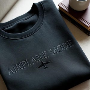 Puede incluir: Sudadera gris oscuro con la frase "AIRPLANE MODE" bordada en el pecho, junto con un peque&ntilde;o gr&aacute;fico de avi&oacute;n. La sudadera est&aacute; doblada cuidadosamente, con un libro y una taza en el fondo.