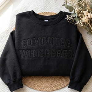 Puede incluir: Sudadera negra de cuello redondo con la inscripción "COMPUTER WHISPERER" bordada en negro. La sudadera es de material suave y se muestra sobre una superficie blanca con flores secas.