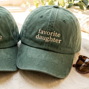 May include: A green baseball cap with the embroidered text "favorite daughter" in white. The cap is made of a soft material and has a curved brim. Sunglasses are visible in the bottom right corner.