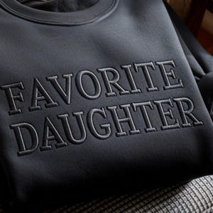 May include: A dark gray sweatshirt with the embroidered text "FAVORITE DAUGHTER" in a lighter gray. The sweatshirt has a crew neck and a soft, textured appearance. The text is centered on the chest.