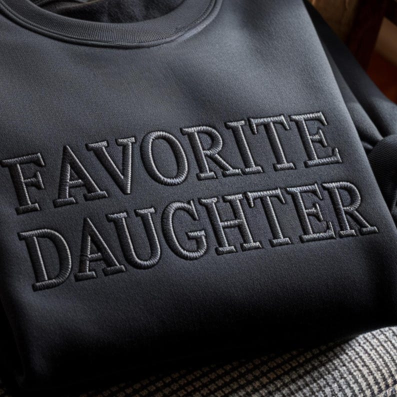 May include: A dark gray sweatshirt with the embroidered text "FAVORITE DAUGHTER" in a lighter gray. The sweatshirt has a crew neck and a soft, textured appearance. The text is centered on the chest.