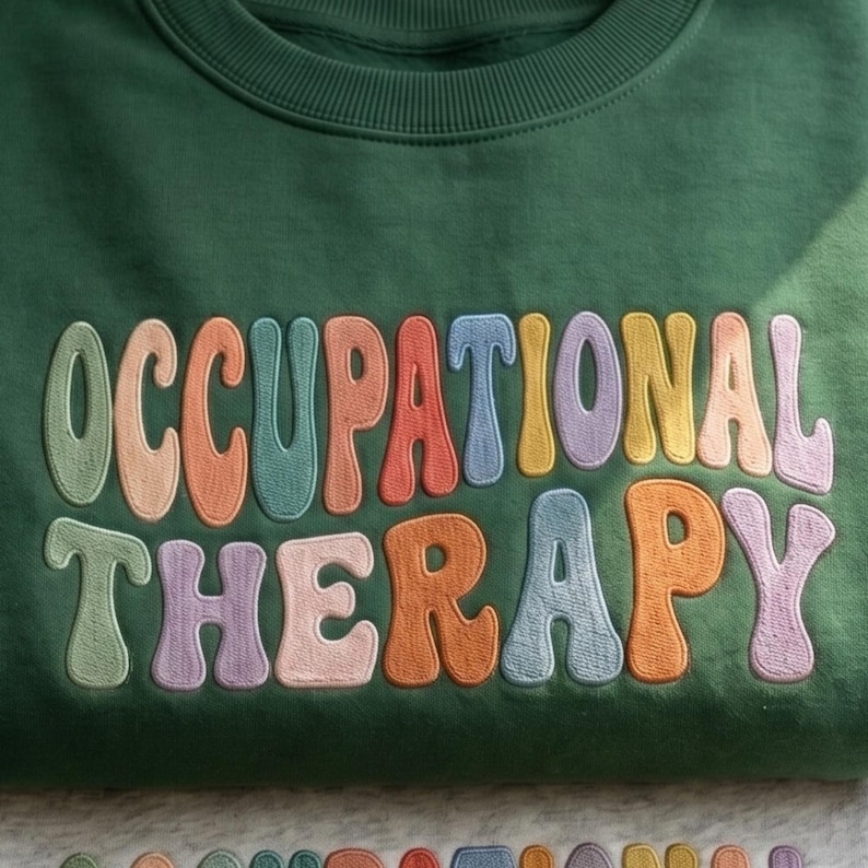 May include: A dark green crewneck sweatshirt featuring the text OCCUPATIONAL THERAPY embroidered in a wavy, multi-colored font. Each letter is stitched in a different pastel or vibrant shade, including light green, peach, orange, blue, and purple.