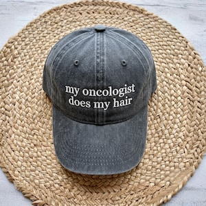 May include: A charcoal gray baseball cap with the embroidered text "my oncologist does my hair" in white. The cap is made of a soft, worn fabric and features a curved brim and adjustable strap. The hat is sitting on a woven, circular mat.