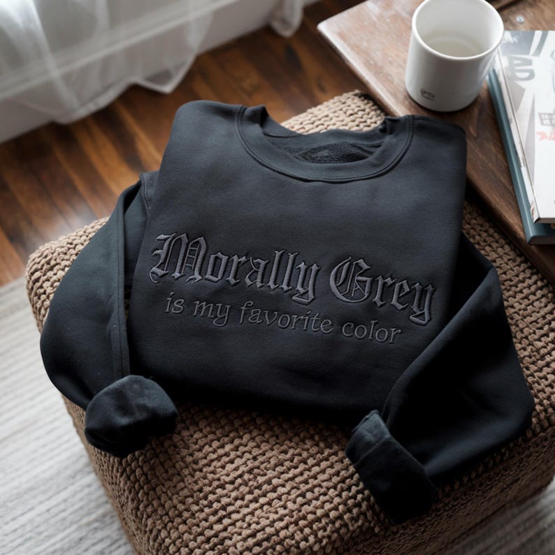Peut inclure: Sweat-shirt noir avec l'inscription brod&eacute;e "Morally Grey is my favorite color". Le sweat-shirt est pli&eacute; sur une surface tiss&eacute;e, avec une tasse blanche et un livre en arri&egrave;re-plan.