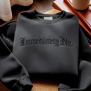 May include: A black sweatshirt with the embroidered text "Immediately No." The crew neck sweatshirt is made of a soft material and has ribbed cuffs and hem. The sweatshirt is folded on a wooden surface.
