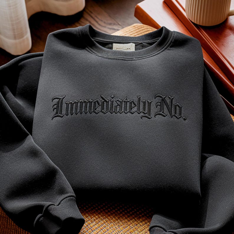 May include: A black sweatshirt with the embroidered text "Immediately No." The crew neck sweatshirt is made of a soft material and has ribbed cuffs and hem. The sweatshirt is folded on a wooden surface.