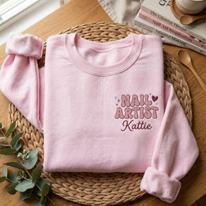 May include: A light pink sweatshirt with the words "NAIL ARTIST Kattie" in a retro font. The sweatshirt has a crew neck and cuffed sleeves. The design includes a heart and star accents.