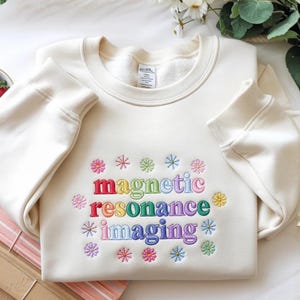 May include: Cream-colored sweatshirt with the words "magnetic resonance imaging" embroidered in colorful letters. The design includes small embroidered flowers in various colors. The sweatshirt has a crew neck and long sleeves.